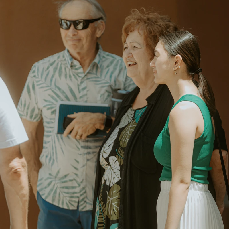 An older man, an older woman, and a young woman stand together in the sun against a brown wall, smiling and looking to the side as if in conversation.