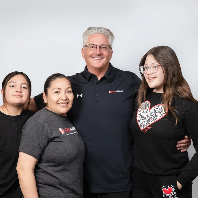 A gray-haired man with glasses smiles, with his arms around a woman and two teenage girls. They are all posing together against a plain white background.