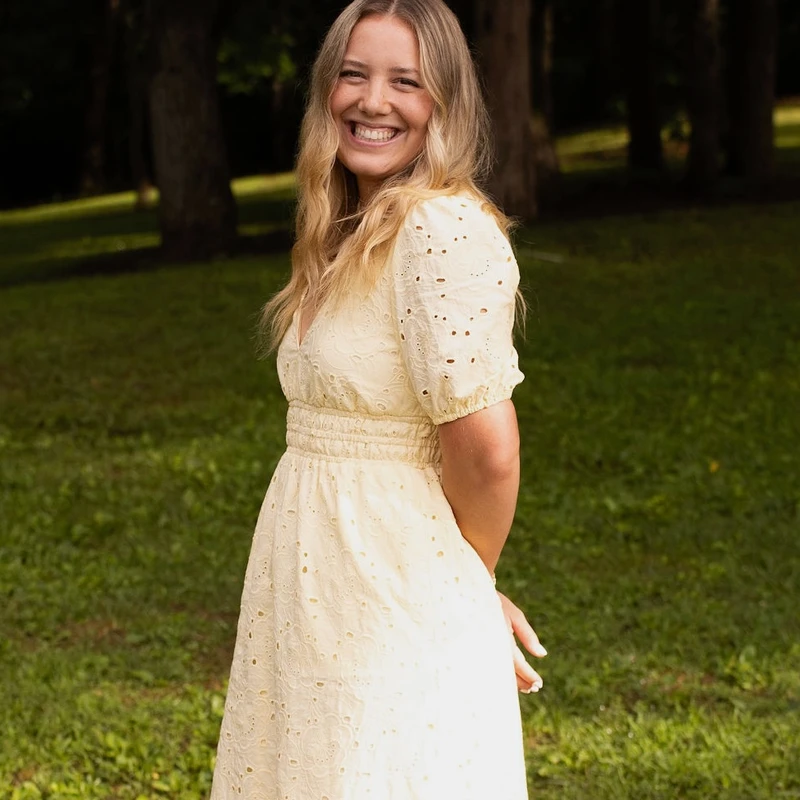 A young woman with wavy blonde hair smiles brightly while standing in a grassy park. She is wearing a pale yellow eyelet dress with puffy sleeves.