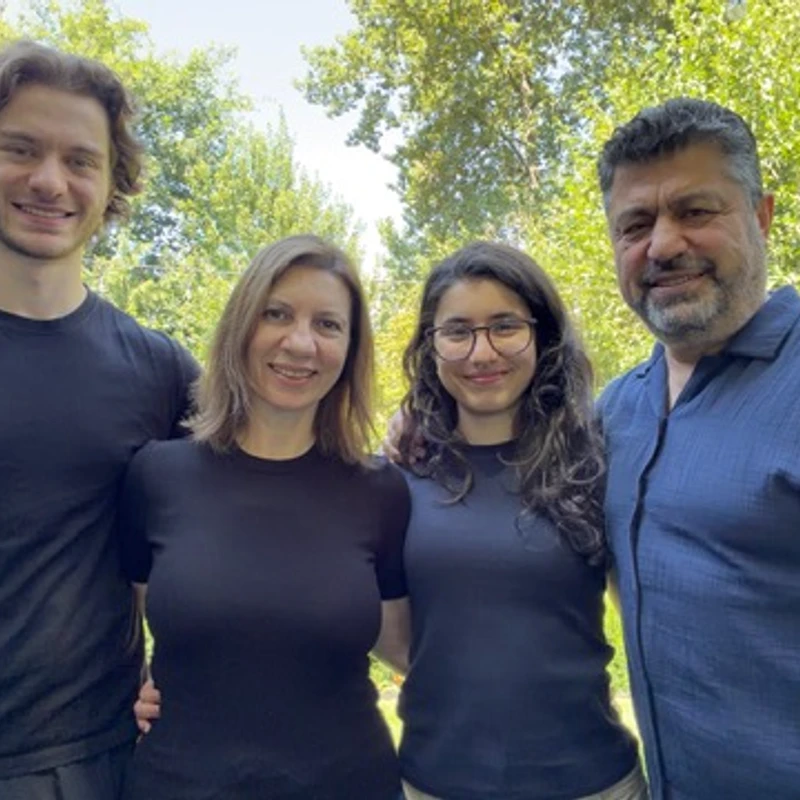 A family of four, including a mother, father, son, and daughter, stand together smiling for a photo in a lush green backyard on a sunny day.