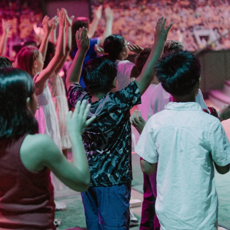 A group of children, viewed from behind, stand with their hands raised high in praise during a worship event in a large venue with colorful lighting.