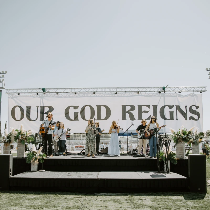A band performs on an outdoor stage under a clear sky, with a large white banner behind them that reads 'OUR GOD REIGNS'.