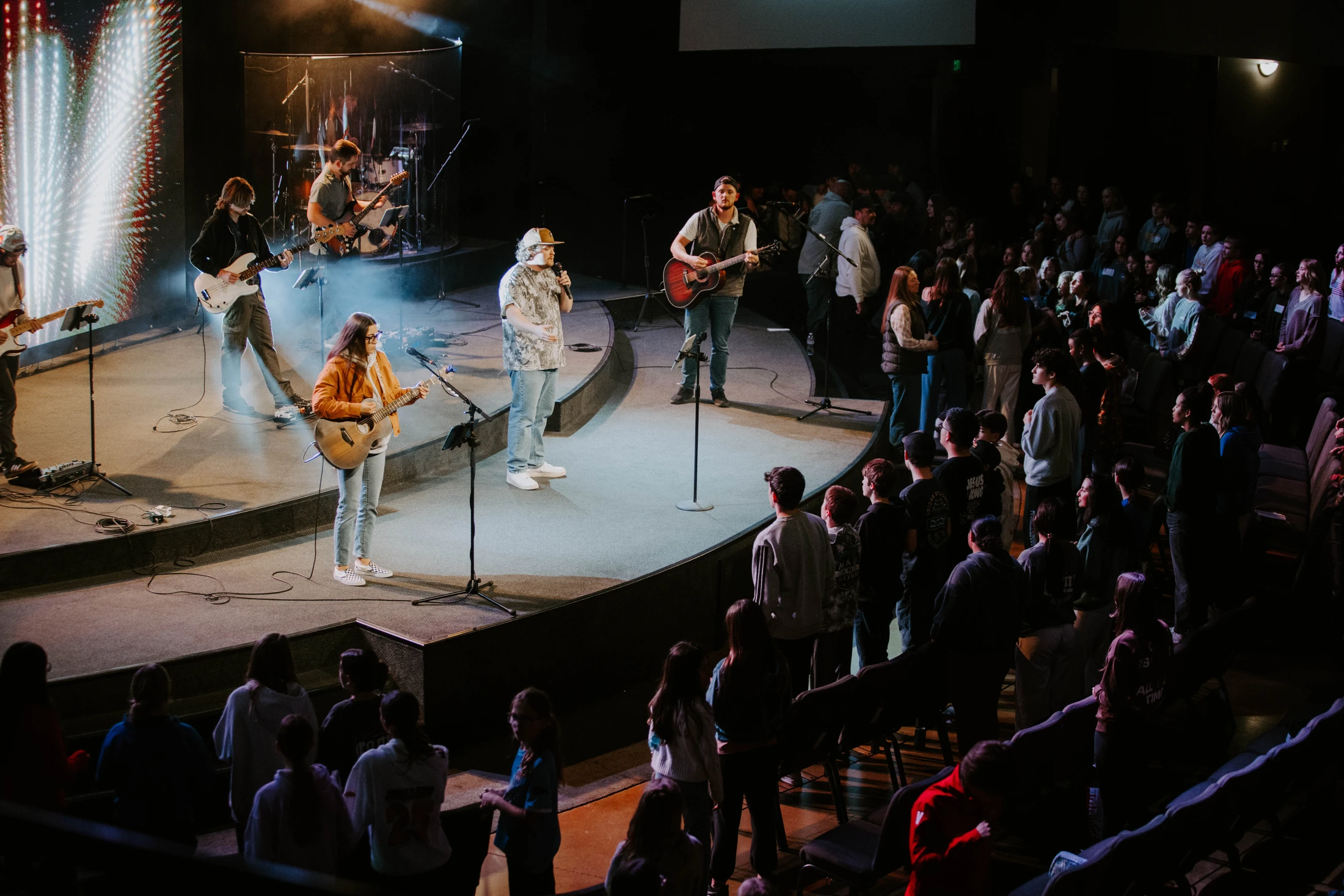 A band performs on a brightly lit stage for a large, standing crowd in a dark auditorium. Musicians play guitars and sing into microphones.