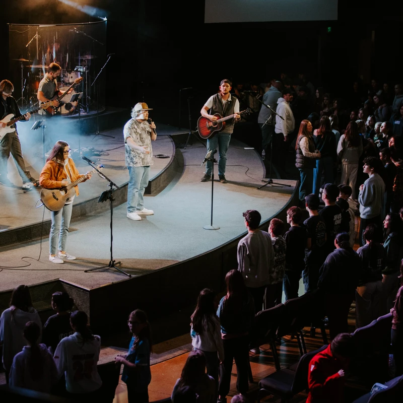 A band performs on a brightly lit stage for a large, standing crowd in a dark auditorium. Musicians play guitars and sing into microphones.