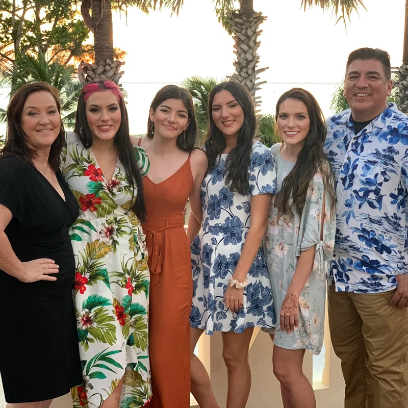 A family of six, two parents and four daughters, smile for a photo on a balcony. Palm trees and a beach at sunset are visible in the background.