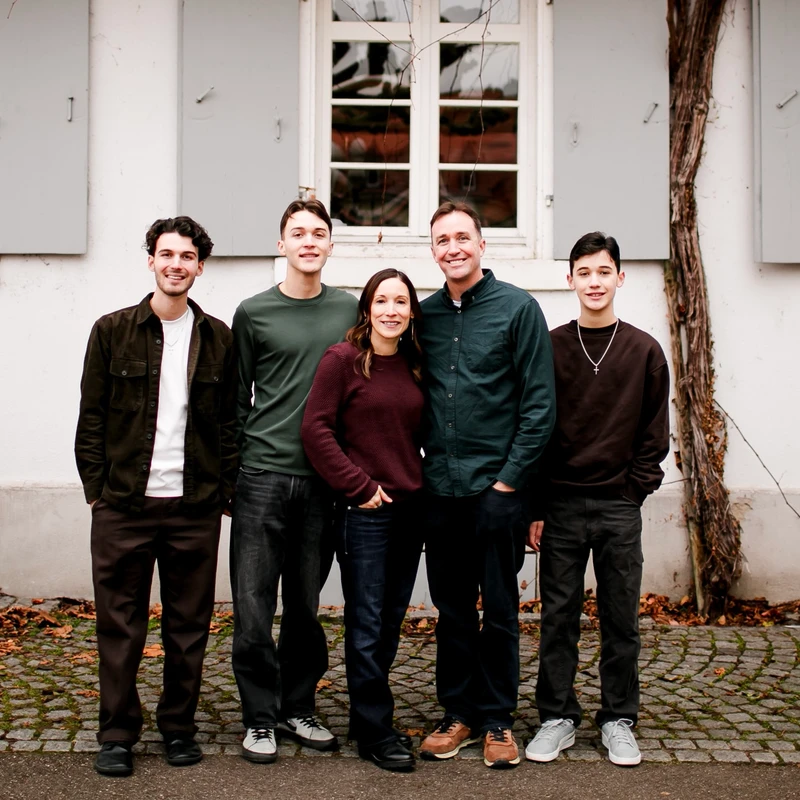 A family of five, two parents and three sons, stand together smiling in front of a white building with grey shutters on a cobblestone path in autumn.