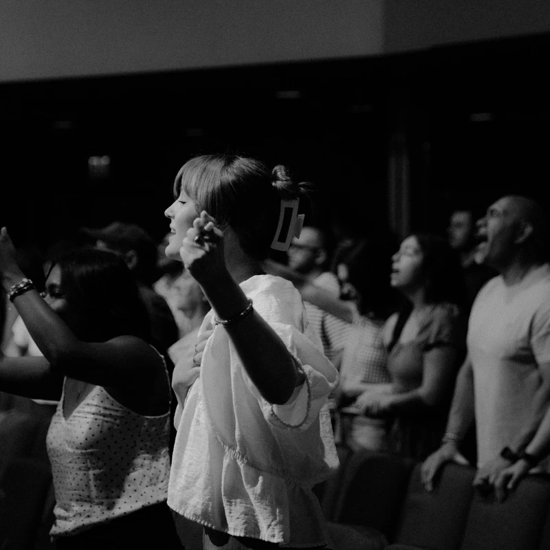 A black and white photo of a group of people in a dark room. They are standing, singing, and some have their hands raised in an act of worship.