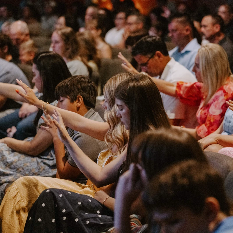 A diverse congregation of people sitting in rows with many individuals raising their hands in an act of worship, with their eyes closed in prayer.