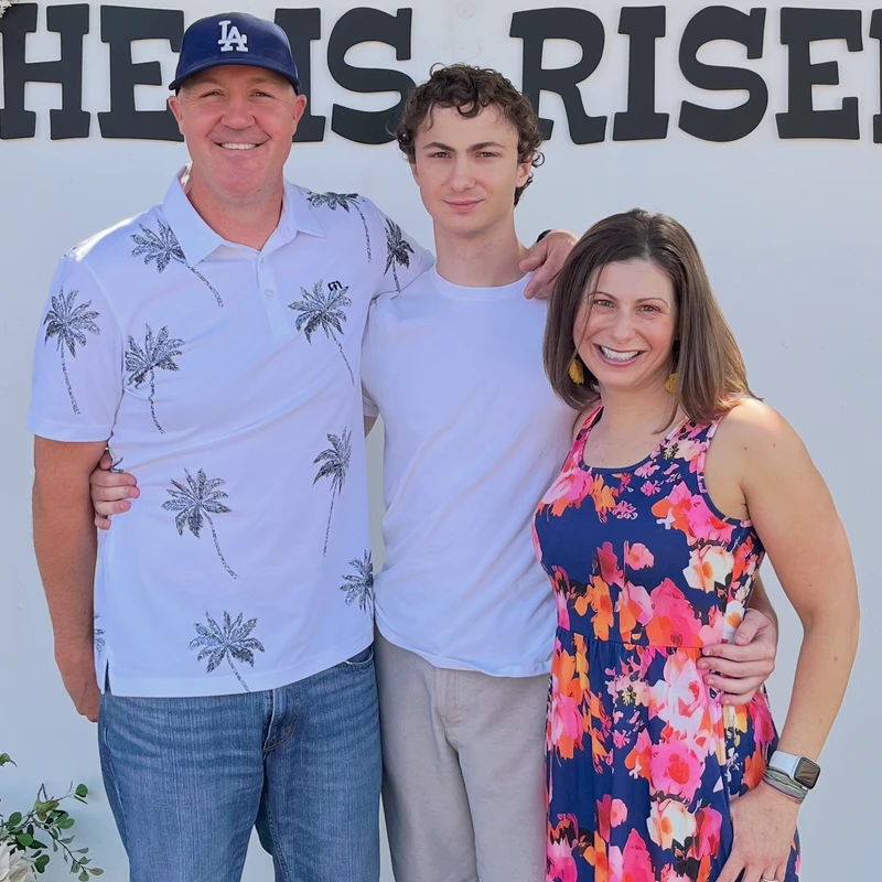A man, teenage boy, and woman stand together and smile for a photo in front of a white wall with large black letters that read 'HE IS RISEN'.