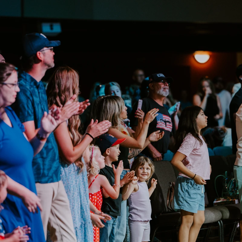 A diverse crowd of adults and children stands together, clapping enthusiastically. One little girl in the foreground smiles at the camera.