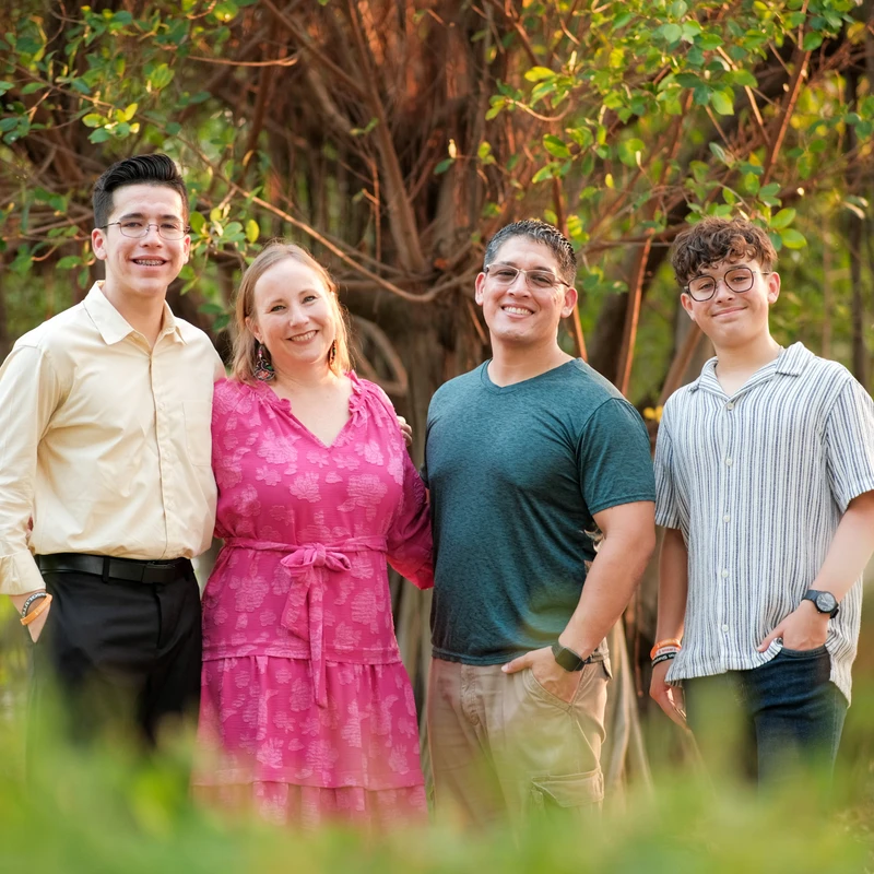 A family of four, two parents and two teenage sons, stand together smiling for a portrait in a park. They are in front of a large tree with warm evening light.