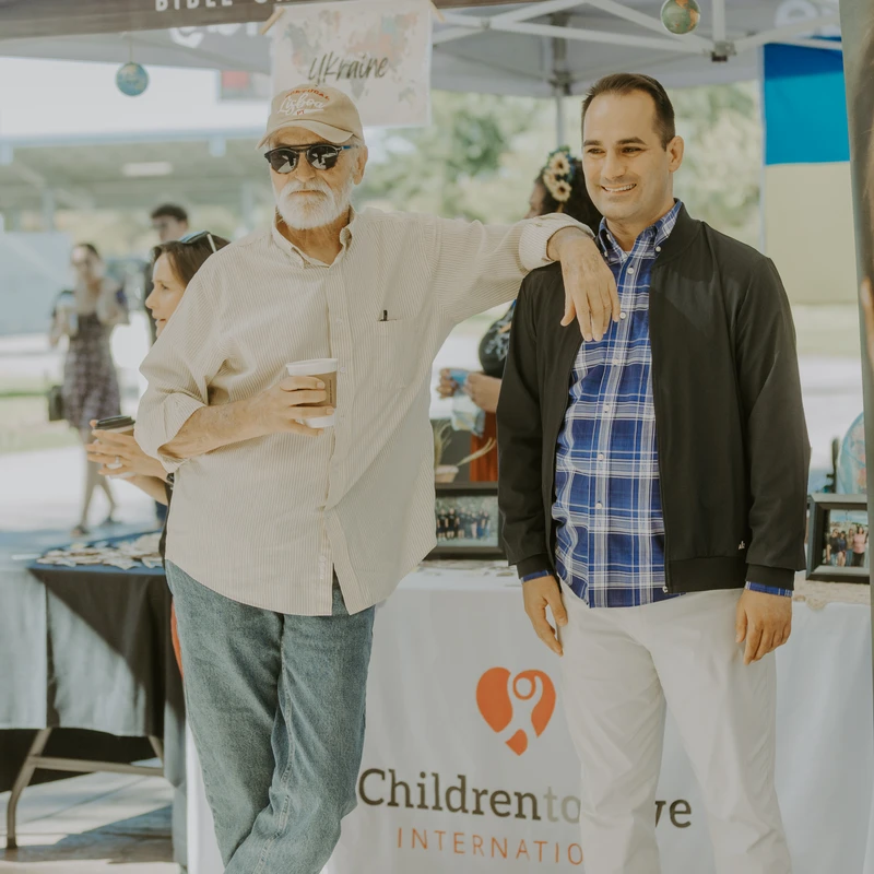 An older man with a white beard and a younger man stand together smiling under a tent for The Bridge Bible Church at an outdoor event.
