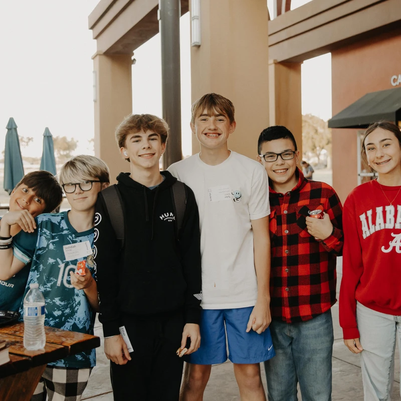 A group of six teenagers, five boys and one girl, smile for the camera while standing together on a patio outside a building with a 'CAFÉ' sign.