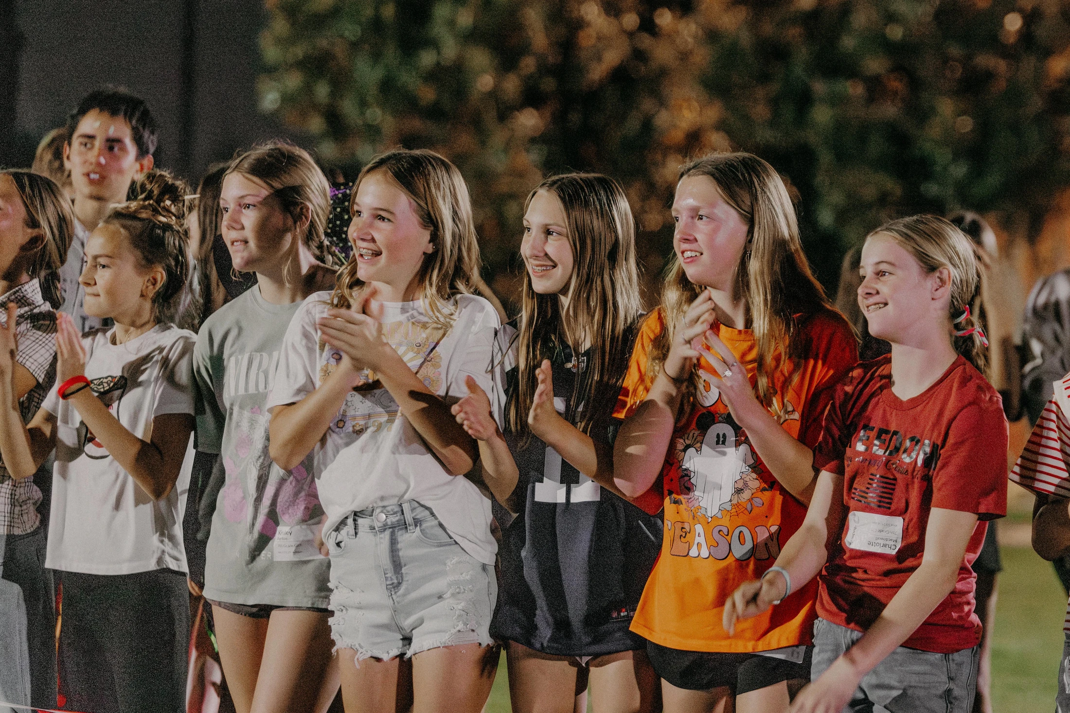 A group of young girls stand in a crowd outdoors at night, smiling and clapping as they watch something off-camera with engaged, happy expressions.