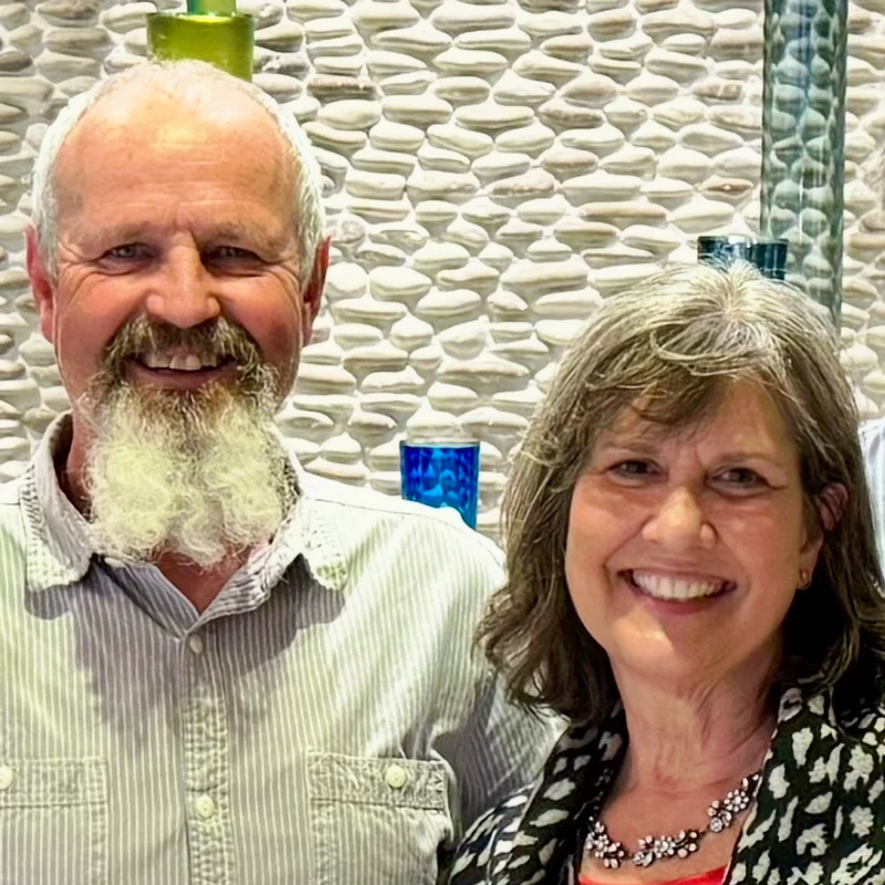A smiling man with a white goatee and a woman with graying hair stand together in front of a textured stone wall, happily posing for a picture.