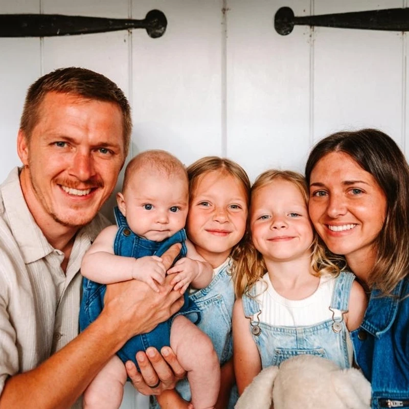 A happy family of five, with a father, mother, two young girls, and a baby, smile for the camera while wearing denim outfits against a white paneled wall.