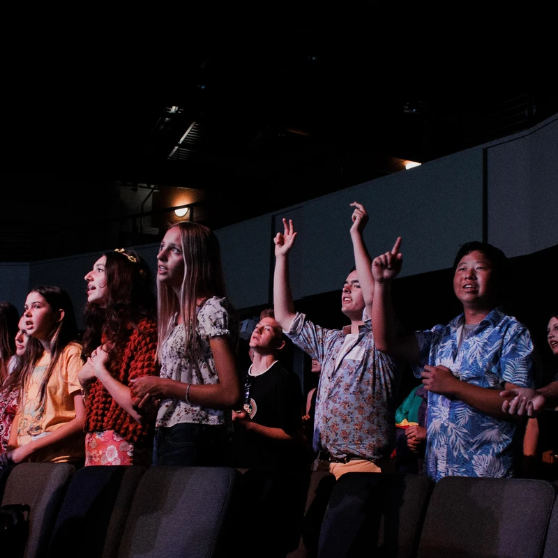 A diverse group of young people stand in a dimly lit auditorium, looking up with engaged expressions. Some have their hands raised in worship or excitement.