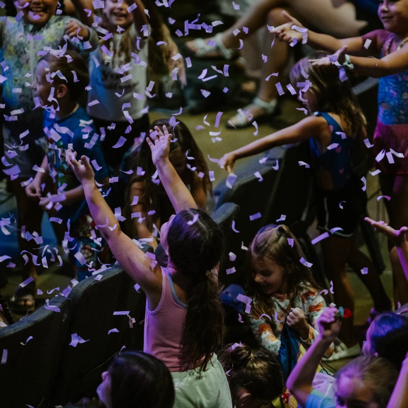A group of happy children with their arms raised in excitement as purple confetti falls all around them during an indoor celebration.