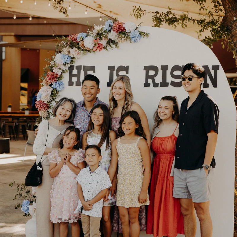 A large, happy, multi-generational family poses for a photo outdoors in front of a white arch decorated with flowers that reads, "HE IS RISEN."
