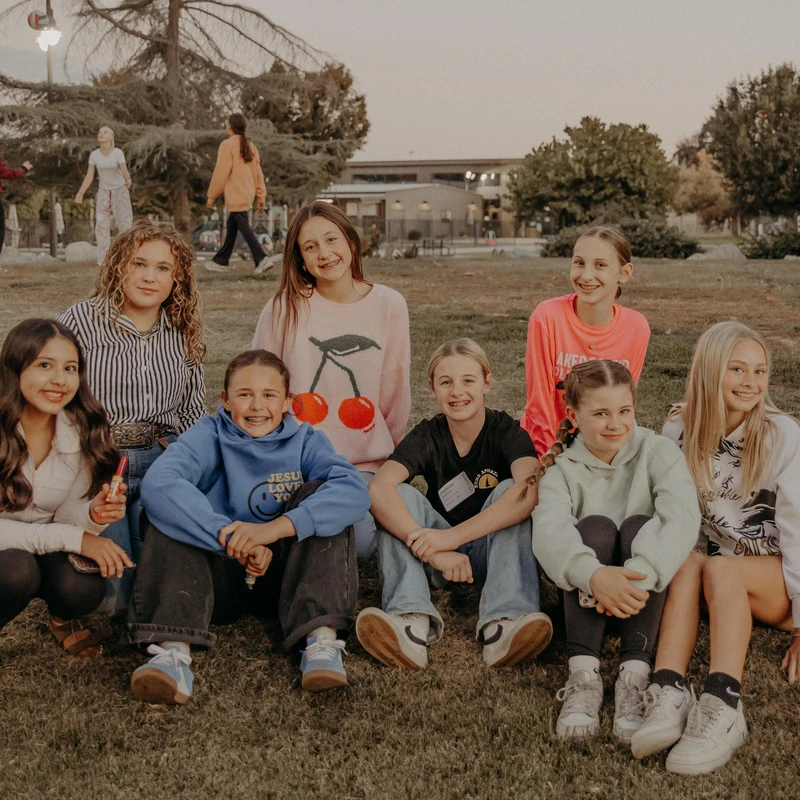 A group of eight young girls in casual clothes sit together on a grassy lawn, smiling and posing for a group photo in a park at dusk.