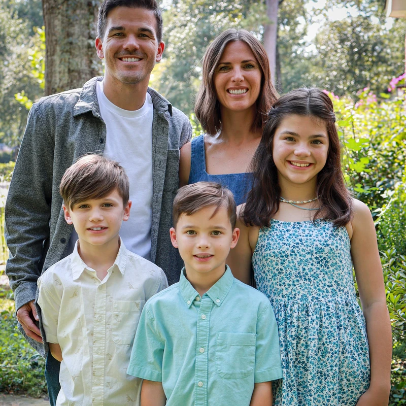A family of five, a mother, father, two sons, and a daughter, smile for a portrait while standing together outdoors in a lush, green garden.