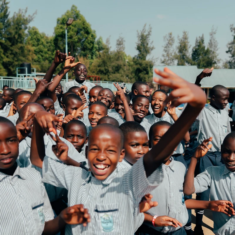 A large group of happy Black schoolchildren in matching striped uniforms smile and make hand gestures for the camera on an outdoor basketball court.