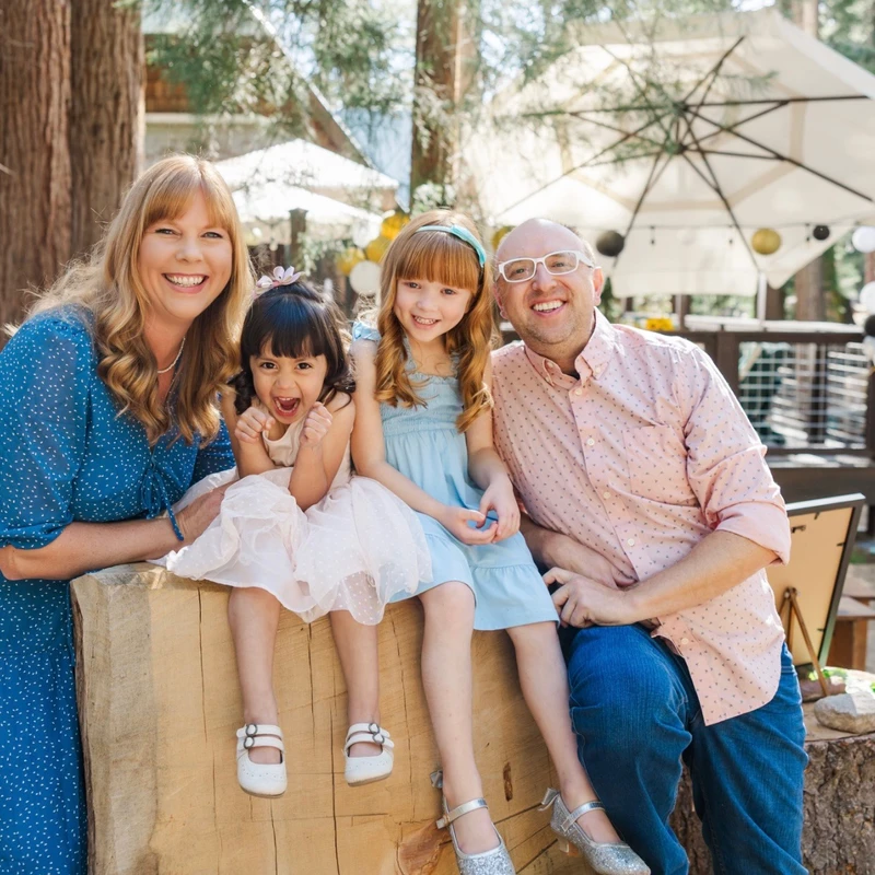 A smiling family of four, including a mother, father, and two young daughters, pose together in a sunny, wooded setting while sitting on a large wooden log.
