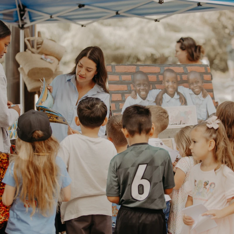 A group of young children gathers around an outdoor stall under a blue tent, listening as a woman shows them a book.