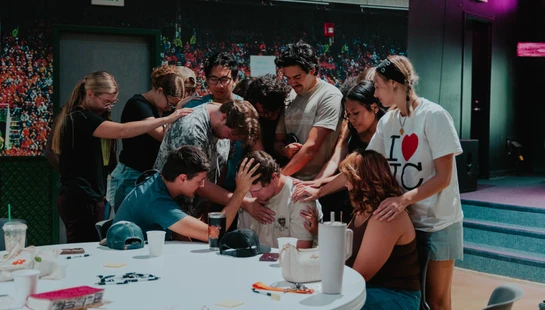 A group of young adults gather around a man seated at a table, placing their hands on him and bowing their heads in a supportive prayer circle.