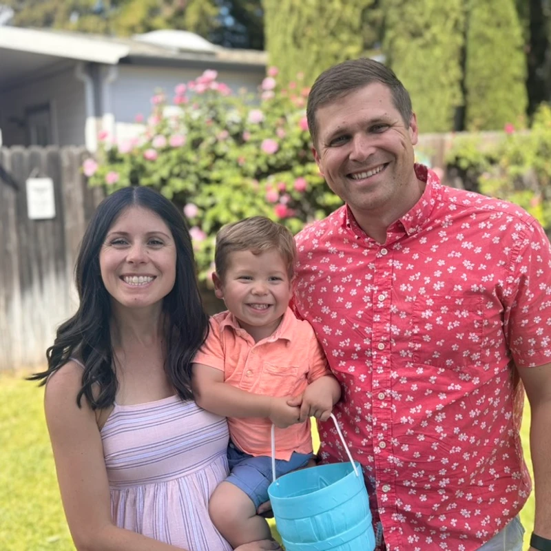 A smiling family of three, a mother, father, and young son, pose for a picture outdoors. The boy sits on his mother's hip and holds a blue plastic basket.