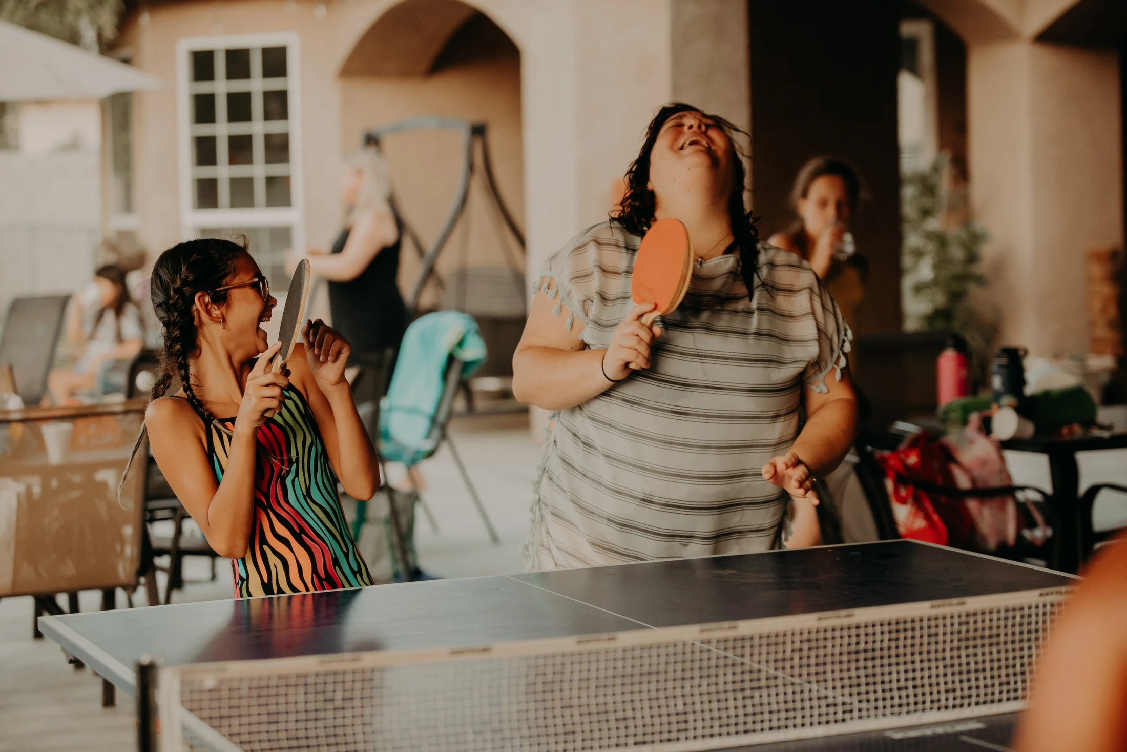 A young girl and a woman laugh joyfully while playing table tennis on a patio, with other people enjoying a gathering in the background.