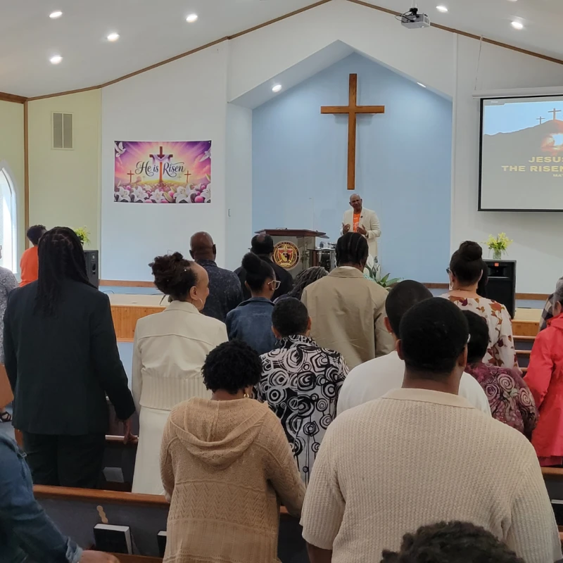 A Black congregation stands in church pews during an Easter service, facing a pastor at the pulpit. A cross and projector screen are visible at the front.