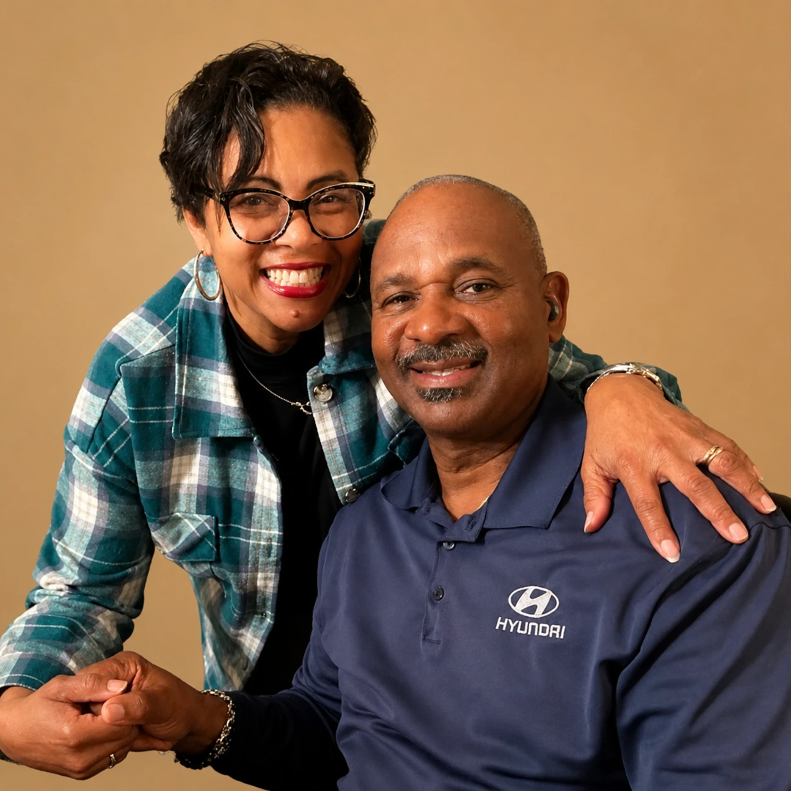 A smiling Black woman in glasses and a plaid shirt stands behind and hugs a smiling Black man wearing a blue polo shirt with a Hyundai logo.