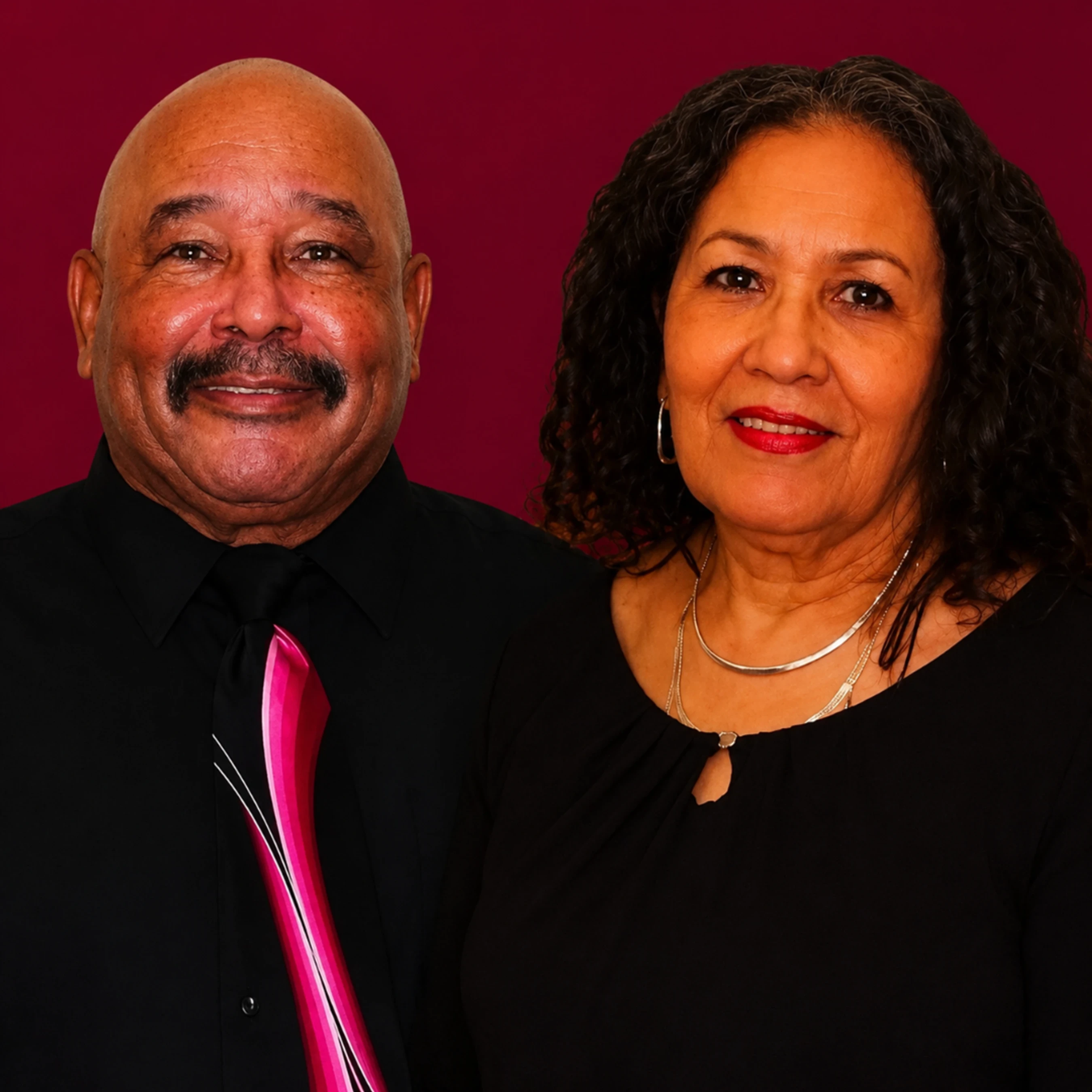 A smiling older couple poses for a studio portrait. The man is bald with a mustache and wears a black shirt with a pink tie. The woman has dark curly hair.