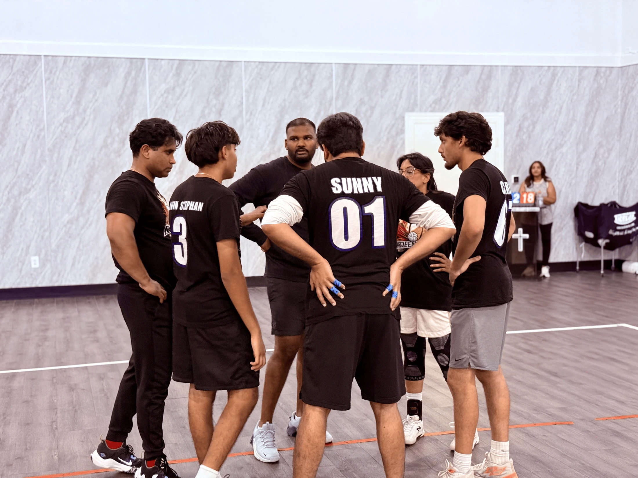 A sports team in black jerseys huddles together on an indoor court while a team leader gives them instructions during a game.