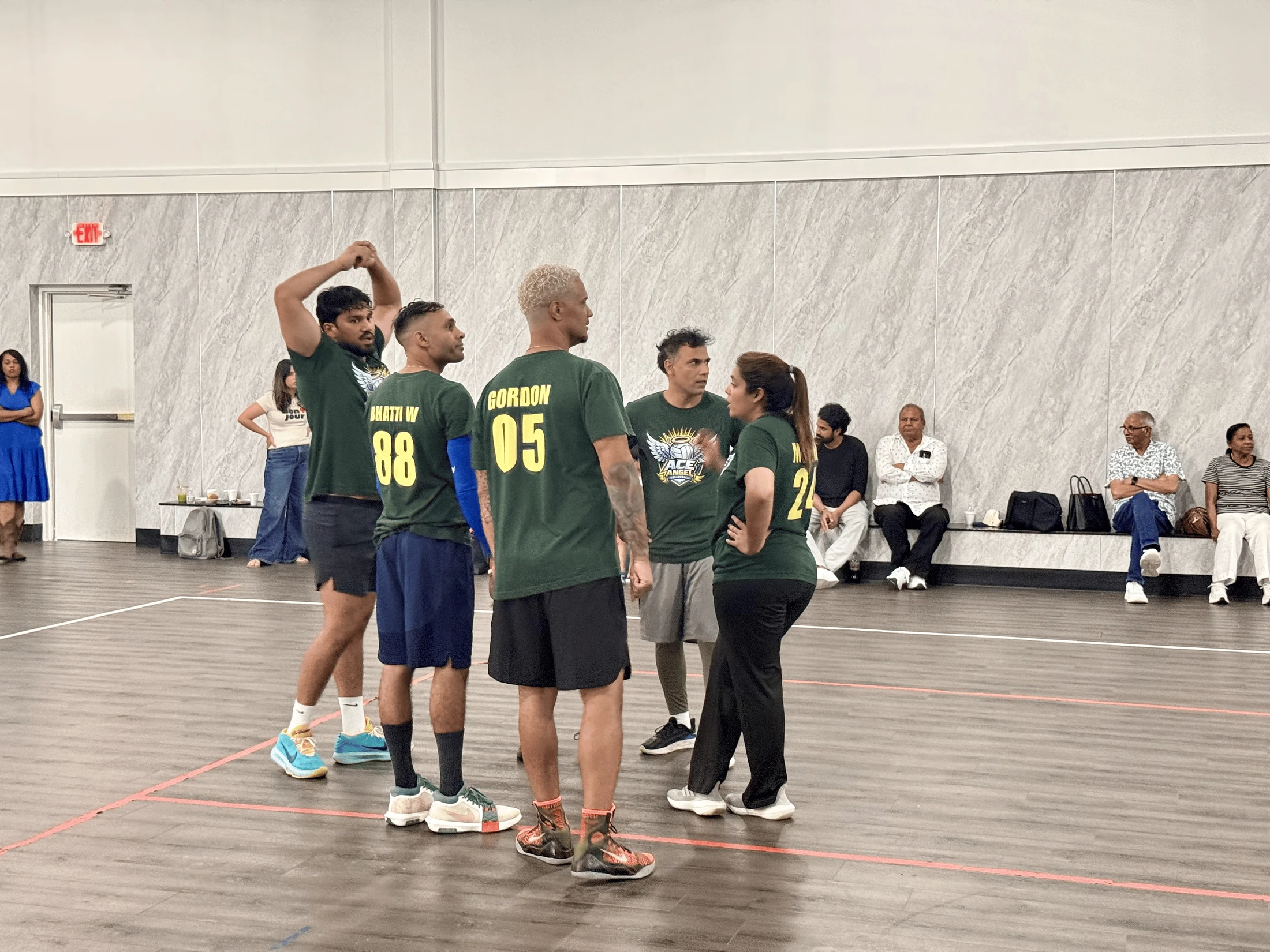 A group of volleyball players in green jerseys stand in a huddle on an indoor court, talking strategy while spectators watch from a bench in the background.