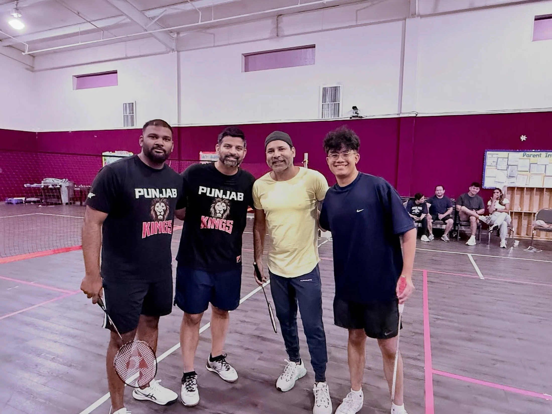 Four men in athletic wear smile for a photo on an indoor badminton court. Two men wear matching black 'Punjab Kings' t-shirts and they all hold rackets.