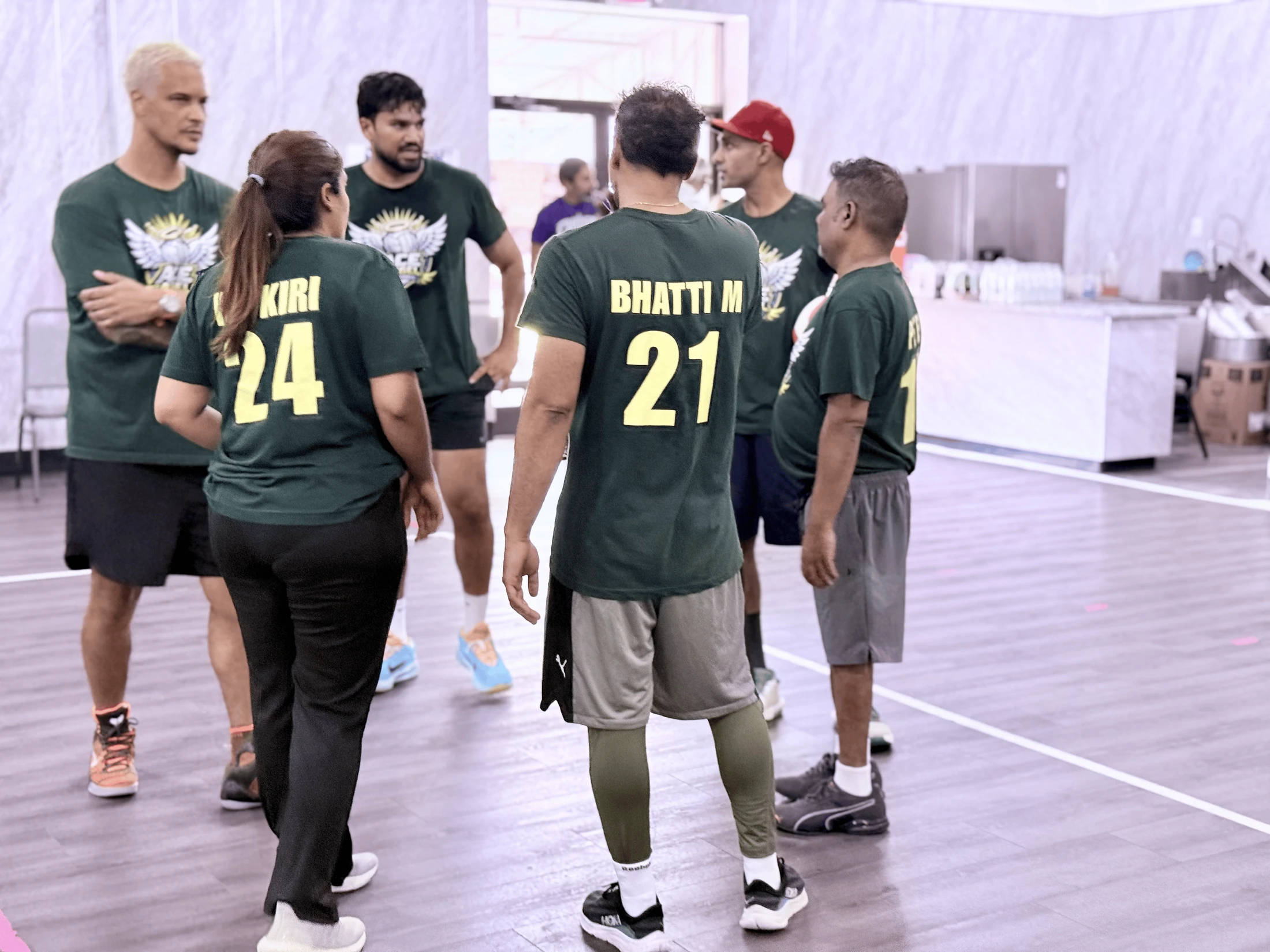 A group of six people in matching green jerseys with yellow numbers huddle together on an indoor court, appearing to talk strategy.