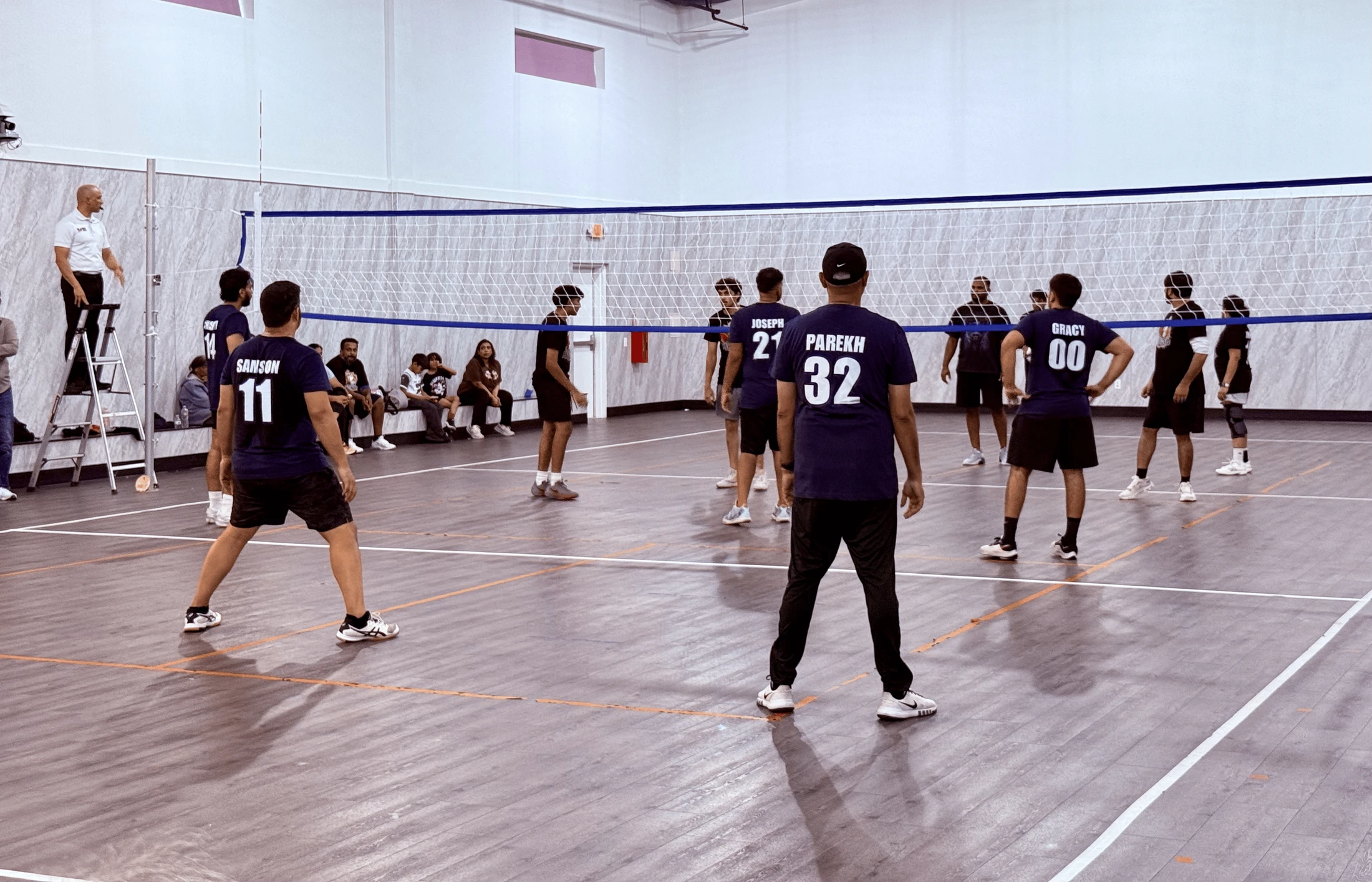 Two teams of men in matching navy blue jerseys face each other across a volleyball net on an indoor court, with a referee watching from a ladder.