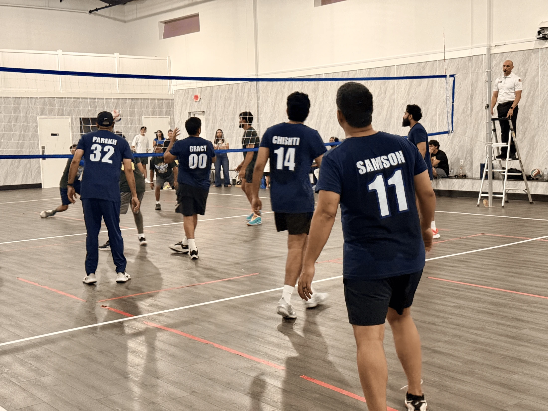 View from behind a men's volleyball team in blue jerseys during a game. A referee watches from a ladder next to the net in an indoor gymnasium.