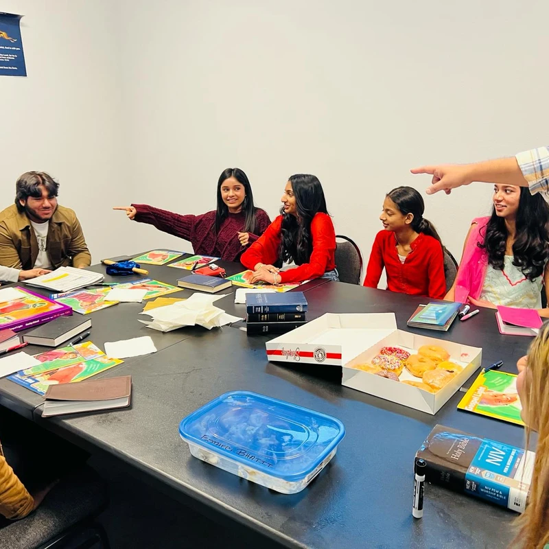 A group of young people sit around a large table with Bibles and donuts, smiling and pointing at each other during a fun and engaging discussion.