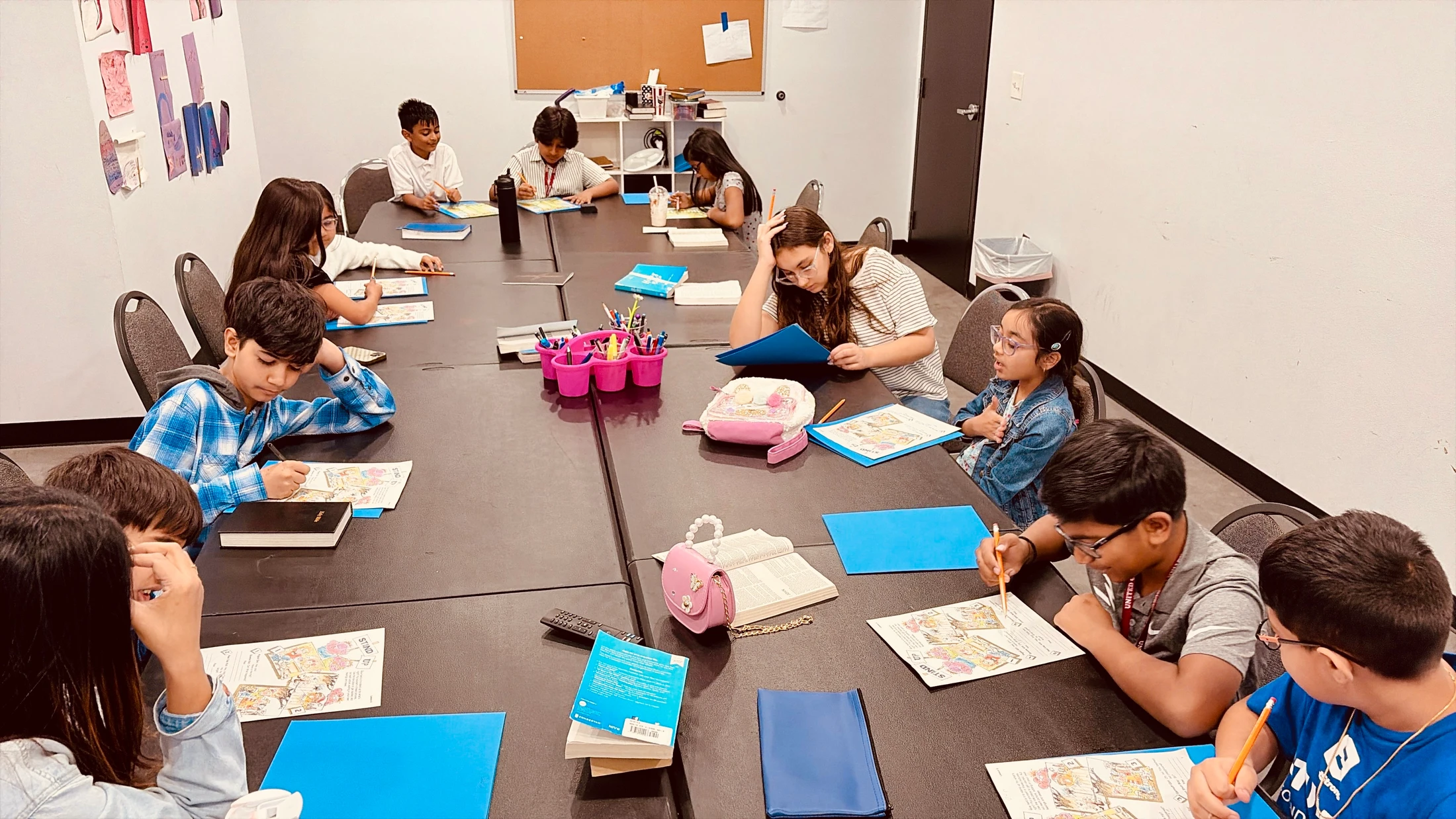 A diverse group of children sit around a long conference table, focused on drawing and writing on worksheets during a class activity.