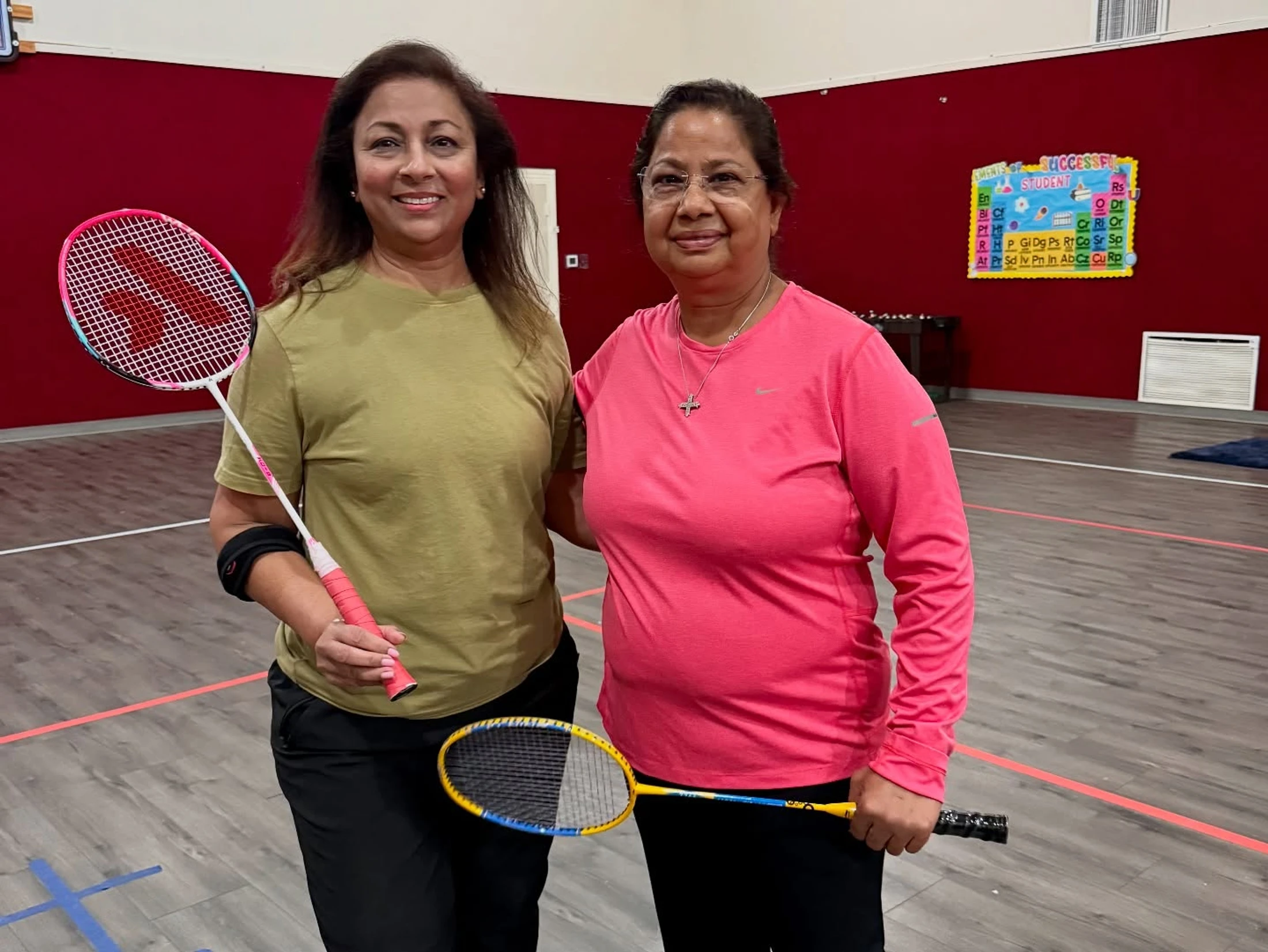 Two middle-aged women in athletic wear stand on an indoor badminton court with a red wall, smiling at the camera and holding their rackets.
