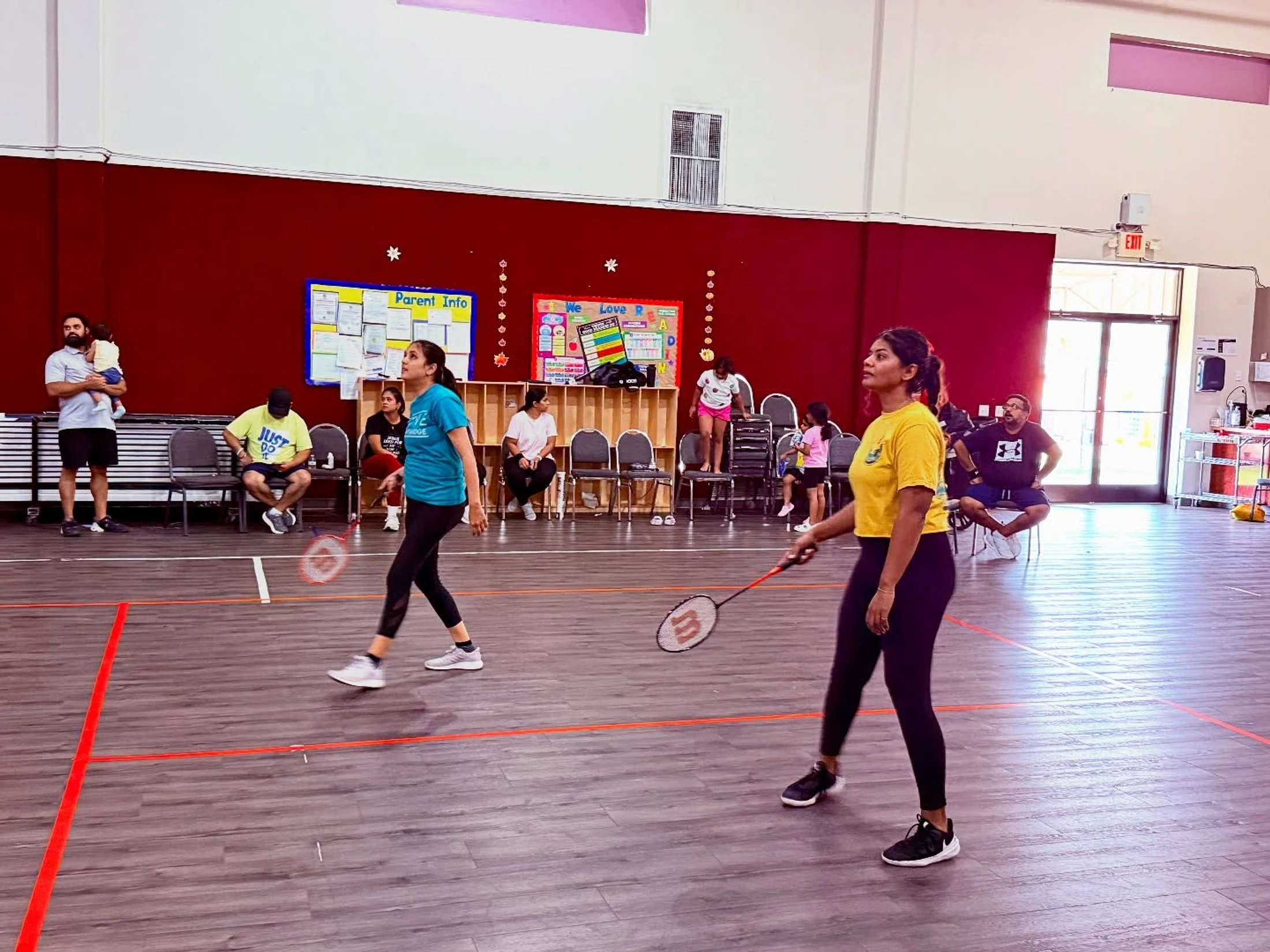 Two women play a game of badminton on an indoor court with red lines, while other adults and children watch from chairs along the wall.
