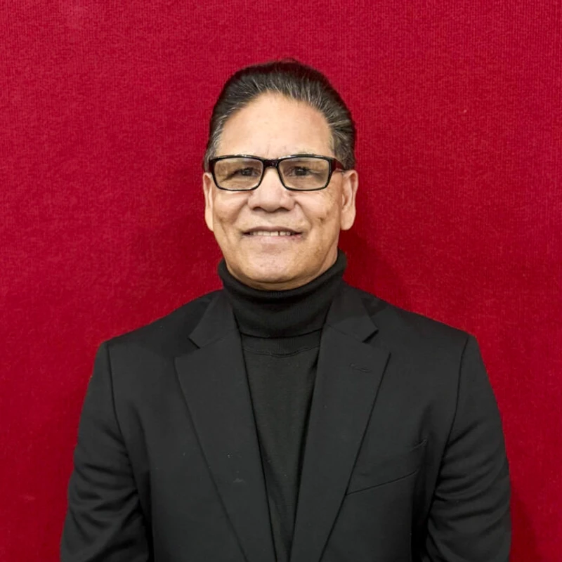 A headshot of a smiling man with dark, slicked-back hair and glasses, wearing a black turtleneck and blazer against a solid red background.