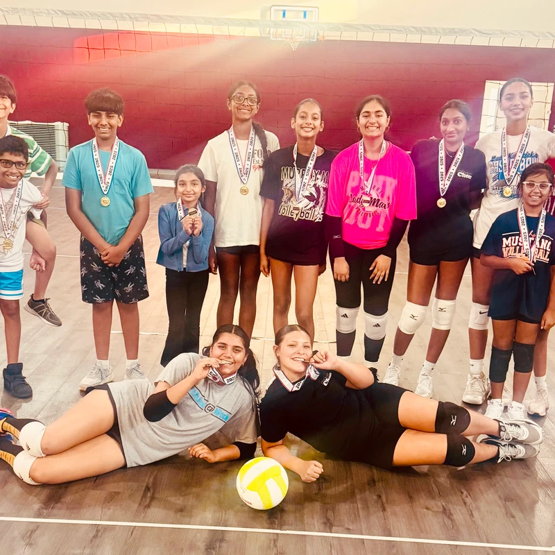 A diverse youth volleyball team poses happily for a group photo in a gym. They are all wearing medals, with two girls lying on the floor with a volleyball.