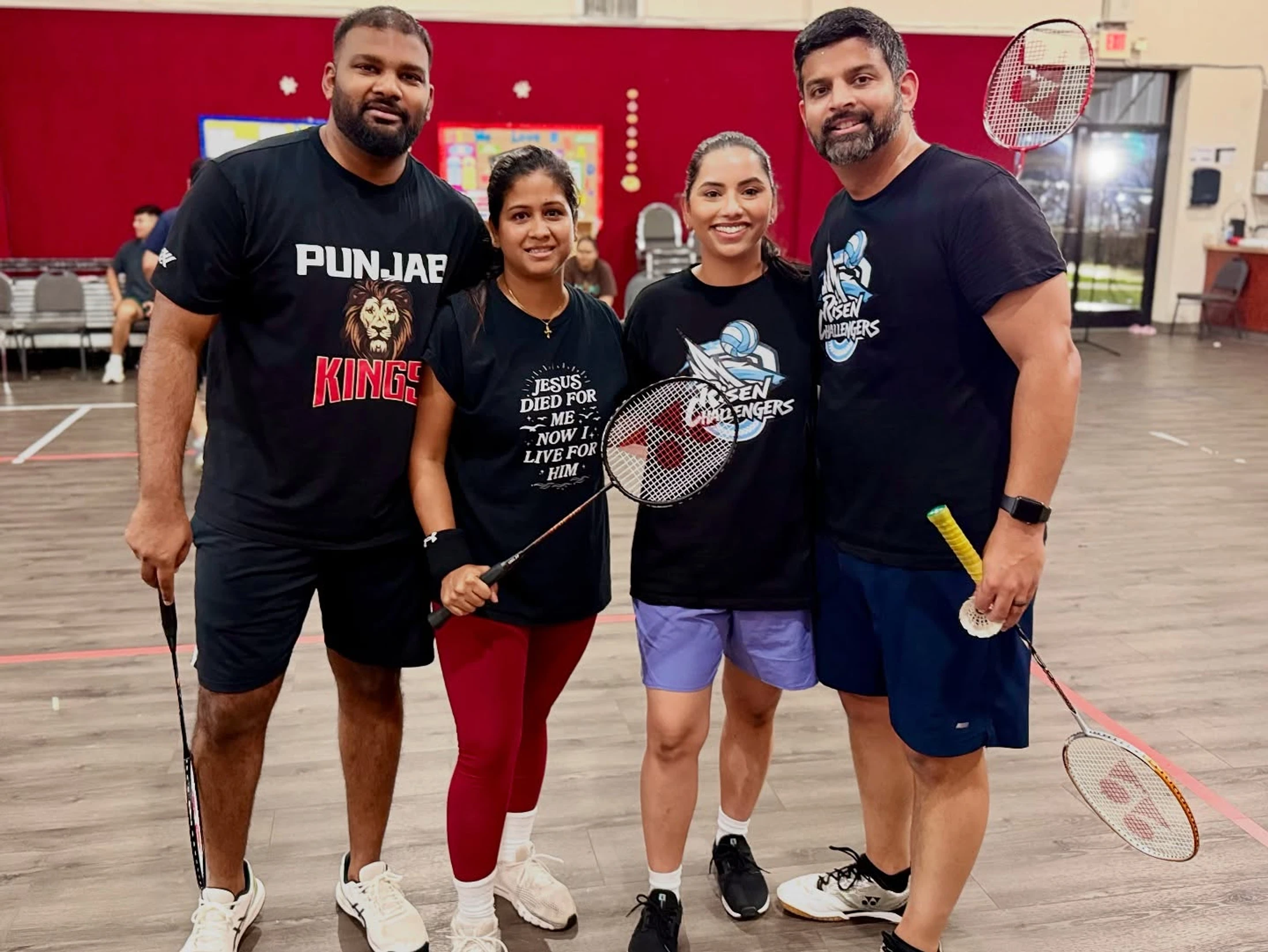 Four friends, two men and two women, in sportswear, smiling and posing with their badminton rackets on an indoor court after a game.