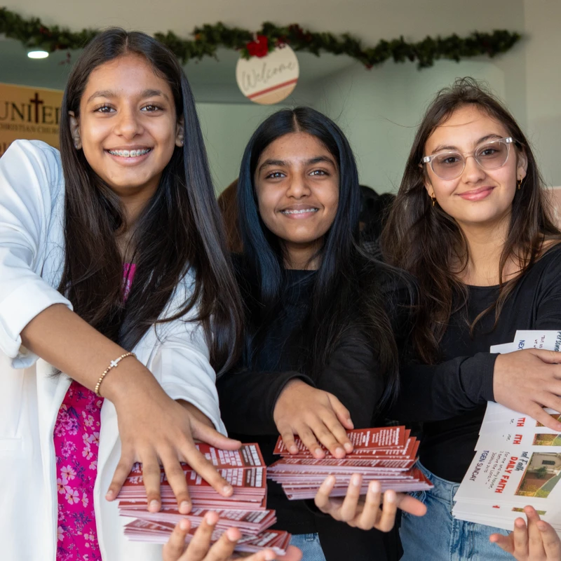 Three smiling young women of South Asian descent stand outside a church, holding stacks of pamphlets to hand out to visitors.