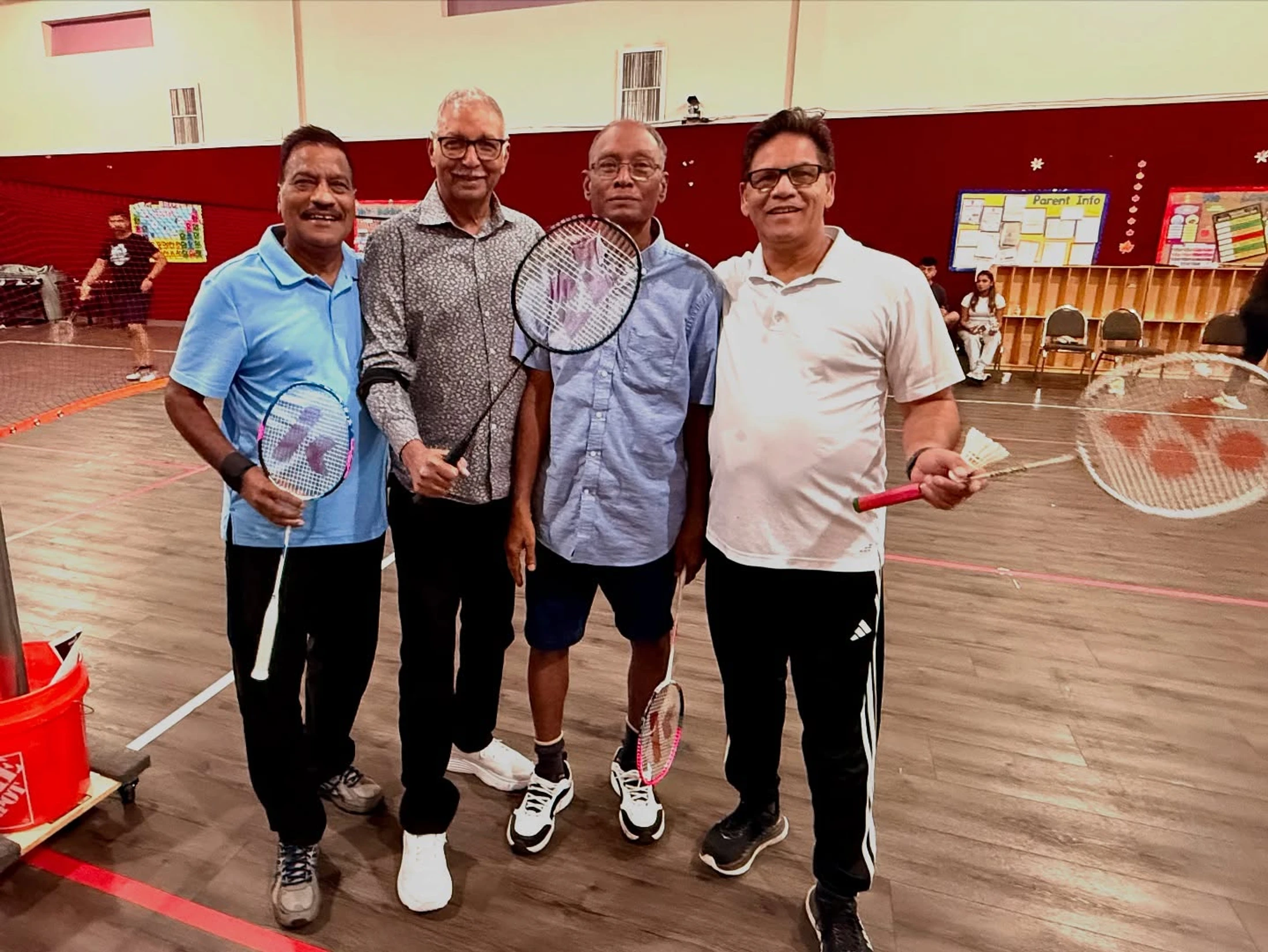 Four smiling older men in sportswear pose for a photo on an indoor badminton court, holding rackets and a shuttlecock after a game.