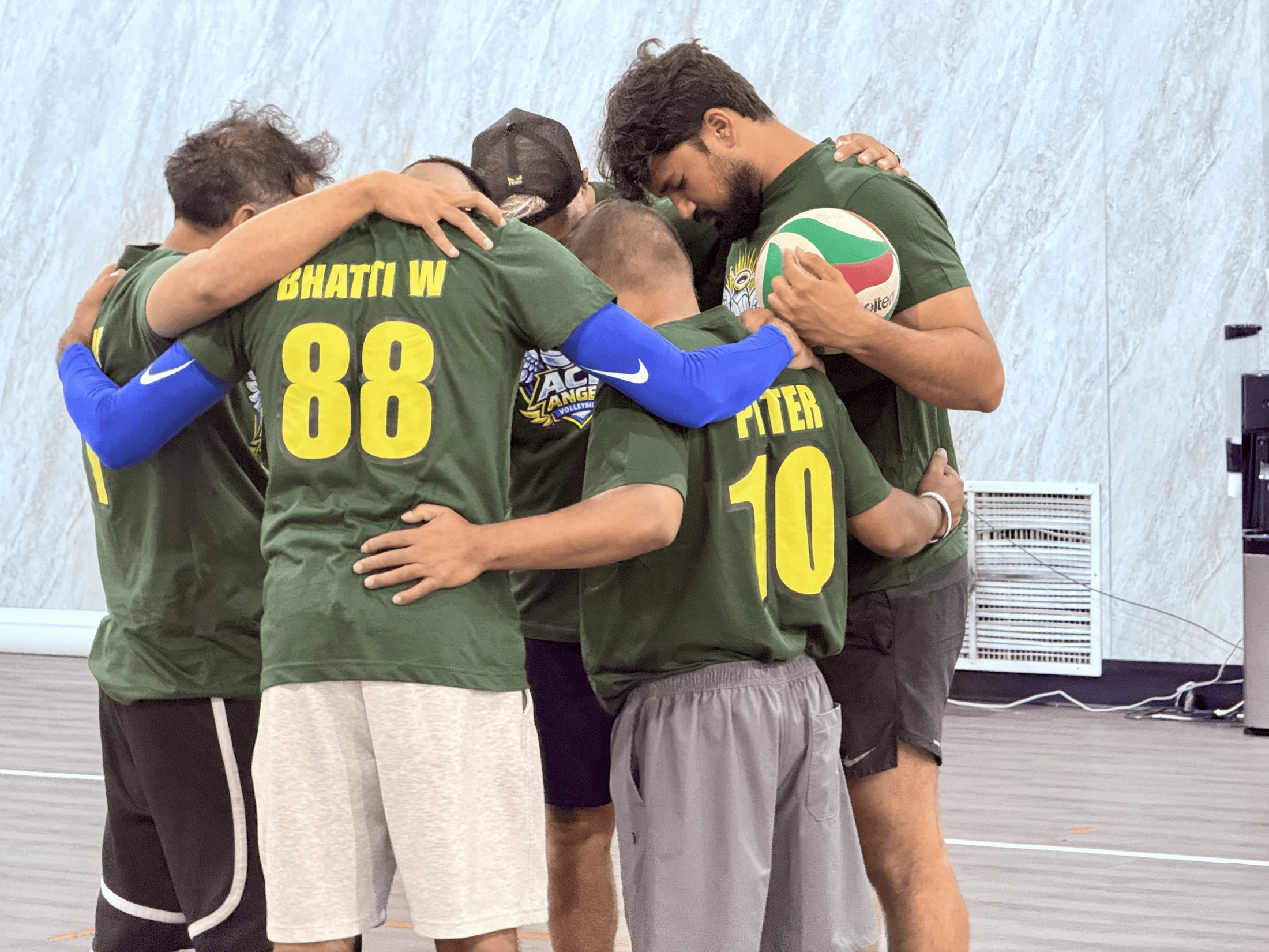 A group of five male volleyball players in green jerseys stand in a tight huddle on the court, arms around each other. One player holds a volleyball.